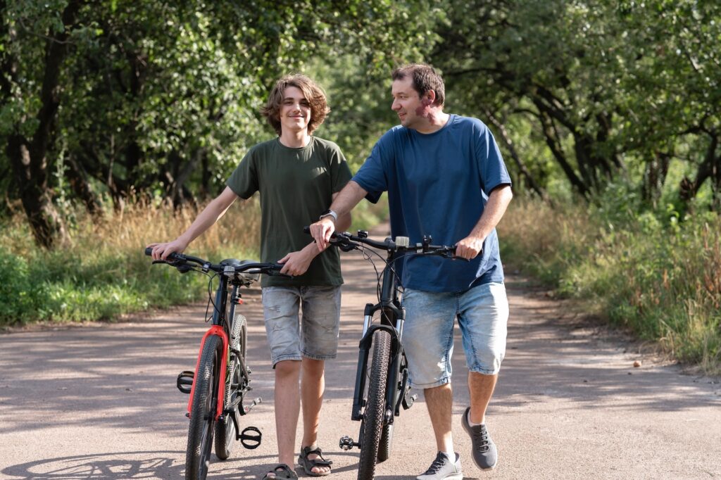 Family outdoor activity, single father and teenager son walking together with bicycles in park at summer, talking and smiling.
